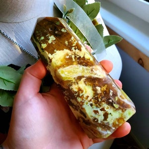 May include: A green and brown polished crystal point with a unique swirling pattern. The crystal is held in a hand.