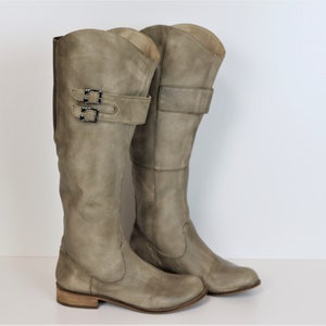 Puede incluir: Un par de botas de cuero gris claro hasta la rodilla. Cada bota presenta un detalle de hebilla en la parte superior de la caña y un tacón bajo apilado. Las botas tienen un aspecto ligeramente desgastado.