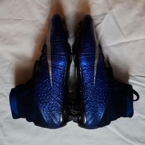 May include: Pair of blue and black athletic shoes with a textured design on the upper. The shoes have a silver swoosh and black laces. The shoes are designed for sports.