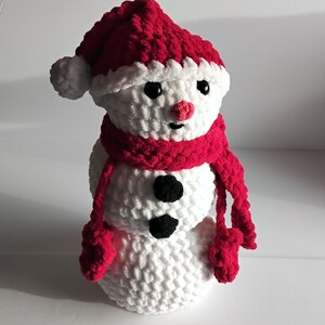 Könnte beinhalten: Ein weißer Häkel-Schneemann mit einer roten Häkelmütze und einem Schal. Der Schneemann hat schwarze Augen, eine rote Nase und zwei schwarze Knöpfe.