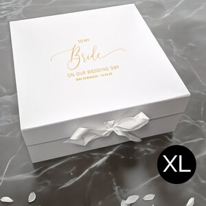 May include: A white gift box with a satin ribbon bow. Gold script on the lid reads "TO MY Bride ON OUR WEDDING DAY" with a date and name. The box is labeled "XL" in a black circle.