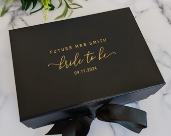 May include: Black gift box with a gold ribbon and the text "Future Mrs. Smith bride to be 09.11.2024" on the top.