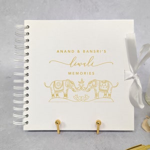May include: A white spiral-bound photo album with gold text and illustrations. The cover reads "ANAND & BANSRI'S Diwali MEMORIES" with two elephant illustrations. A white ribbon ties the album closed. Gold pen and decorative items are in the background.