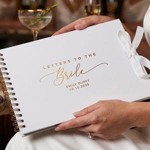 Puede incluir: Un libro blanco con encuadernación en espiral y texto en lámina dorada que dice LETTERS TO THE Bride EMILY DUFFY 10.10.2025. La cubierta presenta una cinta blanca atada en un lazo a la derecha, con una espiral metálica dorada en el borde izquierdo.