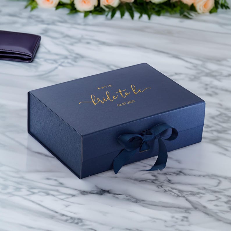 May include: A navy blue gift box with a matching ribbon bow. The box is rectangular and has gold lettering that reads "Katie Bride to be 03.07.2025". The box is on a marble surface with flowers in the background.
