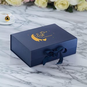 May include: A navy blue gift box with a gold crescent moon and stars design. The box has a gold inscription that reads "Eid Mubarak". The box is tied with a navy blue ribbon.