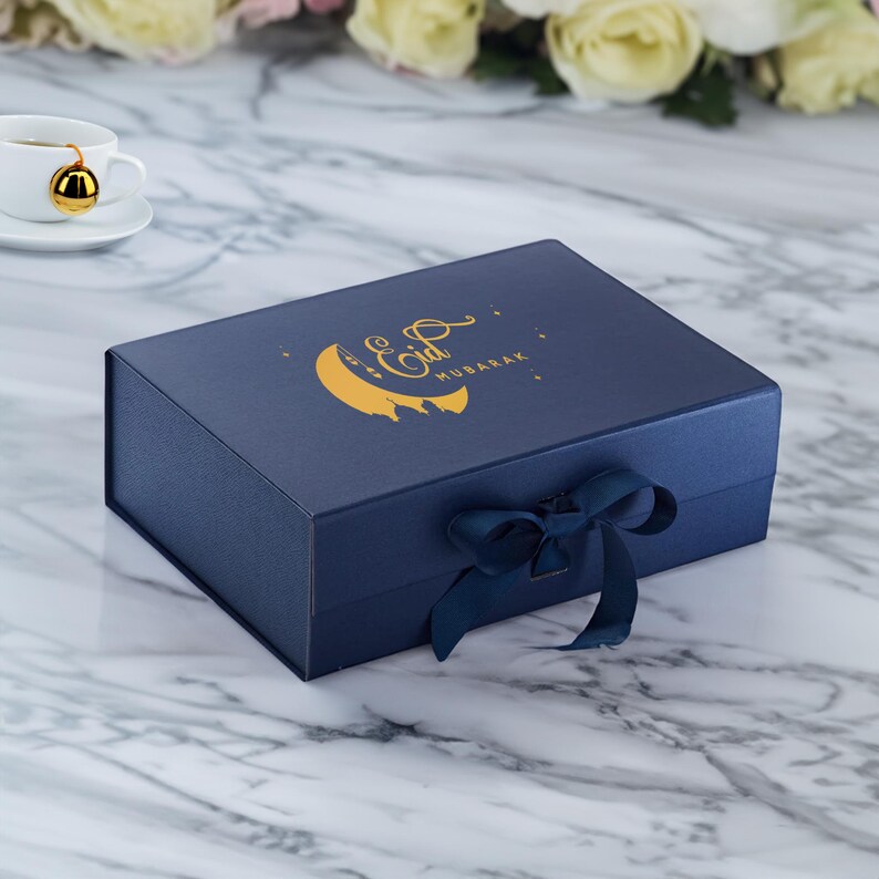 May include: A navy blue gift box with a gold crescent moon and stars design. The box has a gold inscription that reads "Eid Mubarak". The box is tied with a navy blue ribbon.