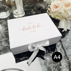 May include: A white gift box with a matching ribbon and bow. The box is labeled "Katie bride to be 03.07.2025" in a copper-colored script. A bouquet of white and peach roses is in the background.