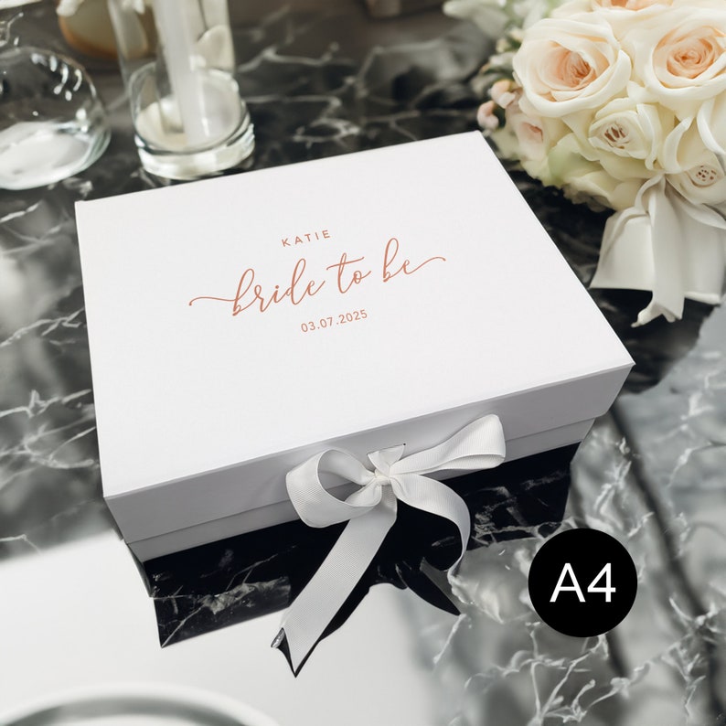 May include: A white gift box with a matching ribbon and bow. The box is labeled "Katie bride to be 03.07.2025" in a copper-colored script. A bouquet of white and peach roses is in the background.