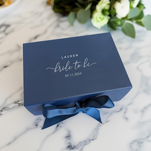 May include: A navy blue gift box with a matching ribbon tied in a bow. The box is inscribed with the name "LAUREN" and the phrase "bride to be" along with the date "09.11.2024" in elegant white script. The box sits on a marble surface.