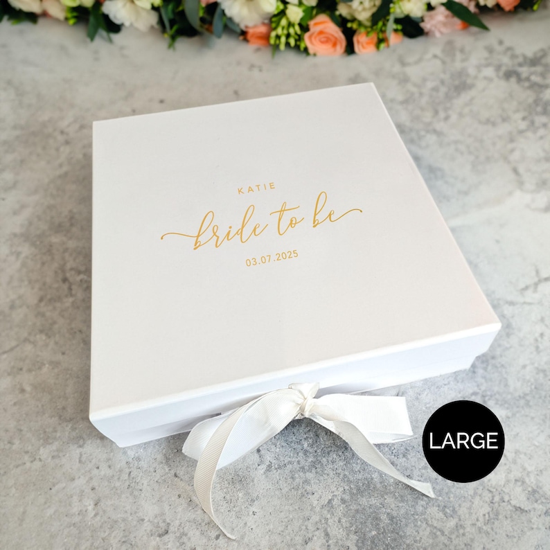 May include: A white gift box with a matching ribbon tied in a bow. The box lid has the words "Katie Bride to be 03.07.2025" in gold script. A black circle with the word "LARGE" is in the bottom right corner.