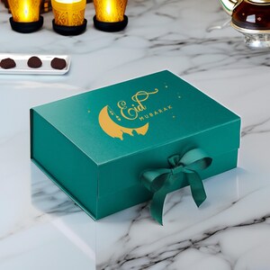 May include: A teal gift box with a gold crescent moon and star design. The box has a gold inscription that reads "Eid Mubarak". The box is tied with a teal ribbon.