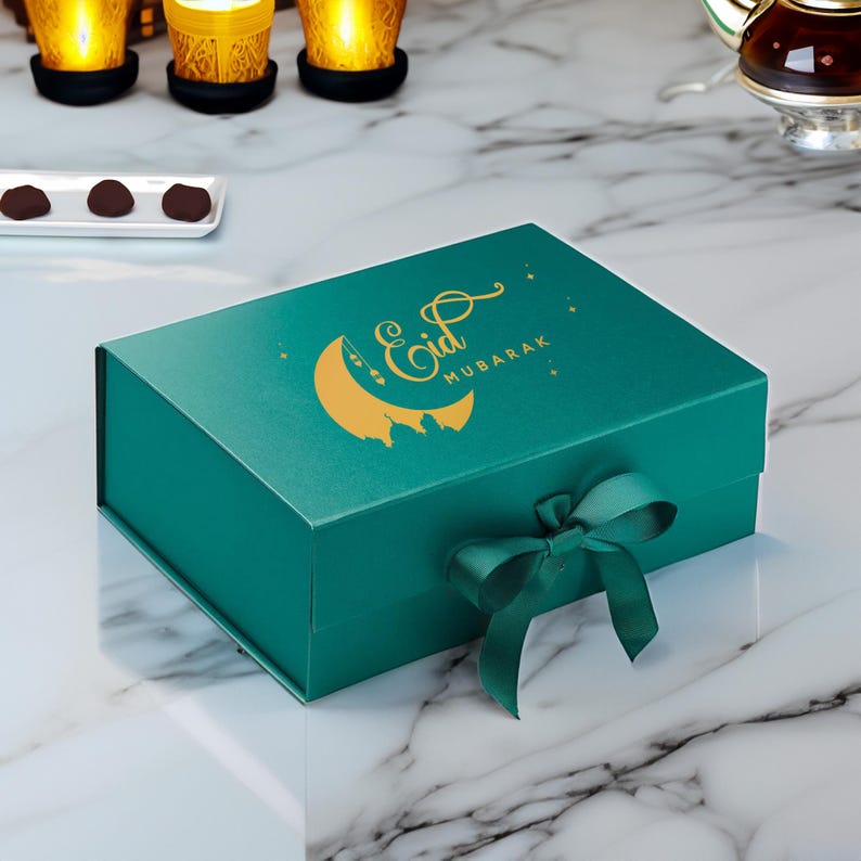 May include: A teal gift box with a gold crescent moon and star design. The box has a gold inscription that reads "Eid Mubarak". The box is tied with a teal ribbon.