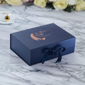 May include: A navy blue gift box with a gold foil crescent moon and the text "Eid Mubarak". The box is tied with a navy blue ribbon.