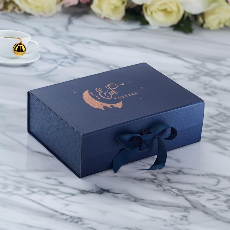 May include: A navy blue gift box with a gold foil crescent moon and the text "Eid Mubarak". The box is tied with a navy blue ribbon.