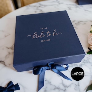 May include: A navy blue gift box with a matching ribbon tied in a bow. The box is labeled "Katie Bride to be 03.07.2025" in a copper-colored script. The word "LARGE" is in a black circle on the bottom right.