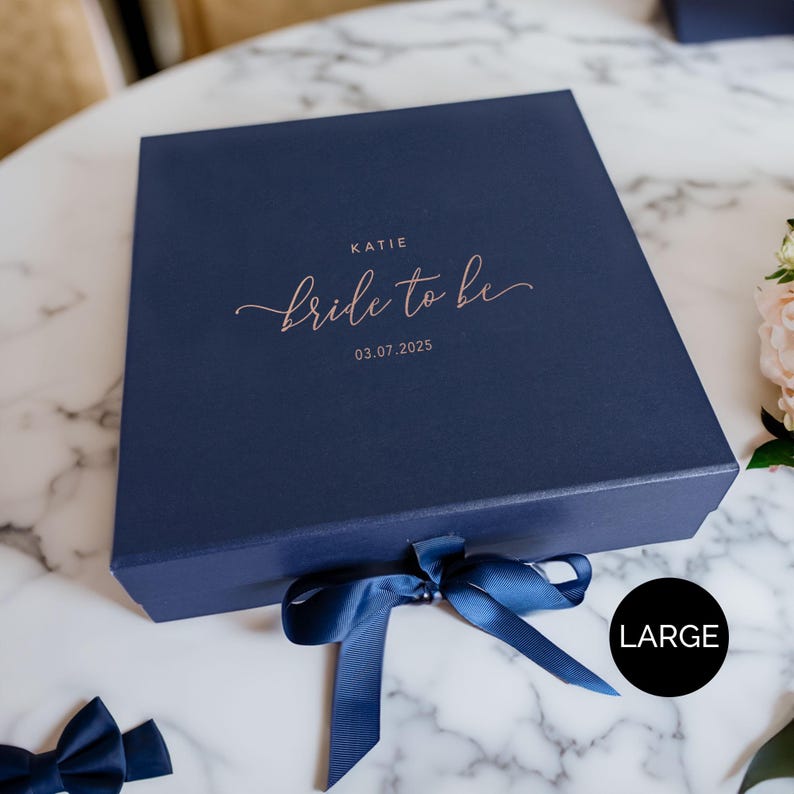 May include: A navy blue gift box with a matching ribbon tied in a bow. The box is labeled "Katie Bride to be 03.07.2025" in a copper-colored script. The word "LARGE" is in a black circle on the bottom right.