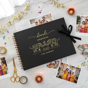 May include: A black Diwali memories photo album with gold spiral binding and a black ribbon. The cover features gold lettering and elephant illustrations. Family photos, decorative tape, scissors, and a small candle are arranged around the album.