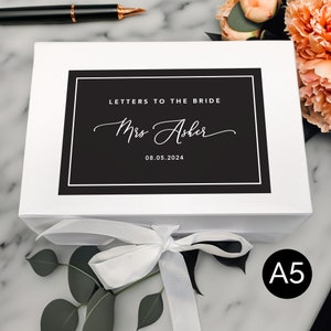 May include: White box with a black label that says "Letters to the Bride" and "Mrs. Asher" with the date 08.05.2024. The box is tied with a white satin ribbon.
