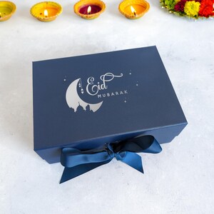 May include: A navy blue gift box with a silver crescent moon and stars design. The box has a silver inscription that reads "Eid Mubarak". The box is tied with a blue ribbon.