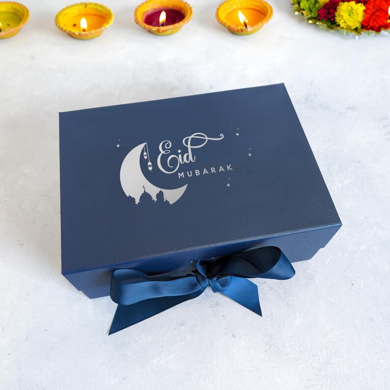 May include: A navy blue gift box with a silver crescent moon and stars design. The box has a silver inscription that reads "Eid Mubarak". The box is tied with a blue ribbon.