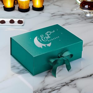 May include: A teal gift box with a silver crescent moon and the words "Eid Mubarak" printed on the top. The box is tied with a teal ribbon.