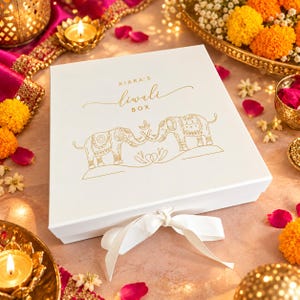 May include: A square white gift box featuring gold foil line art of two elephants and a small diya lamp. The text reads KIARA'S Diwali BOX in gold lettering. A white satin ribbon is tied in a bow at the front center of the box lid.
