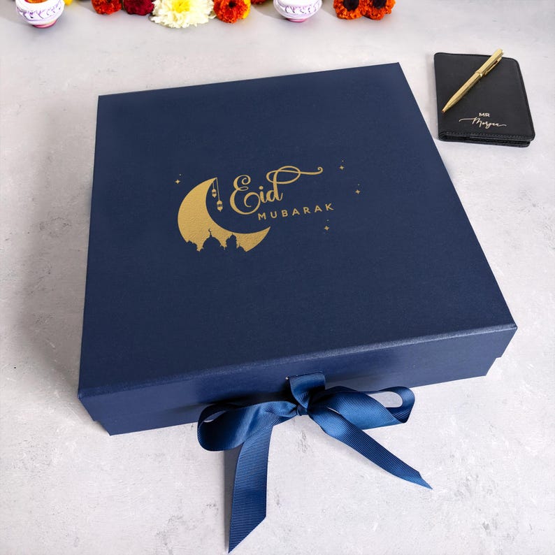 May include: A navy blue gift box with a gold crescent moon and the text "Eid Mubarak". The box is tied with a blue ribbon.