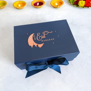 May include: A navy blue gift box with a white ribbon and a gold crescent moon with the text "Eid Mubarak" printed on the top.
