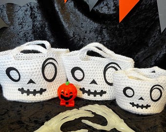 May include: Three white crochet baskets with black skeleton face designs, a small plastic jack-o'-lantern, and a pair of glowing skeleton hands are arranged on a black velvet surface. Orange and gray triangle banners are in the background.