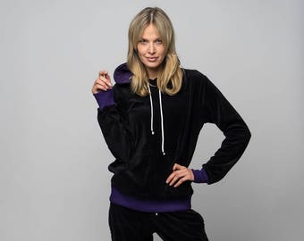 Könnte beinhalten: Ein schwarzer Velours-Trainingsanzug mit violettem Besatz. Der Hoodie hat einen weißen Kordelzug. Das Model trägt den Trainingsanzug und steht auf einem weißen Hintergrund.