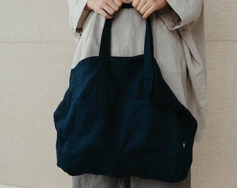May include: A person is holding a dark blue tote bag with a single pocket on the front. The bag is made of a thick, textured fabric.
