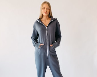 May include: A dark gray jumpsuit with a hood and a white zipper. The jumpsuit has long sleeves, pockets, and elastic cuffs at the ankles. The person is wearing white sneakers. The outfit is casual and comfortable.