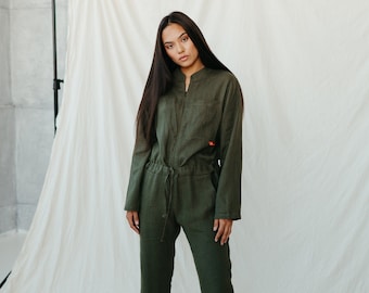May include: A woman wearing a dark green jumpsuit with a zipper and a drawstring waist. The jumpsuit has long sleeves and a relaxed fit.