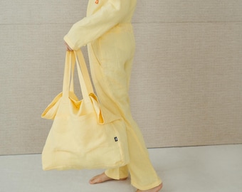 May include: A woman wearing a yellow linen jumpsuit holds a matching yellow tote bag. The bag has a knotted handle and a small black logo.