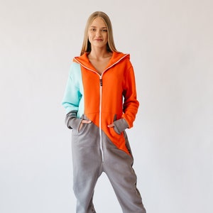 May include: A color-blocked jumpsuit in orange, teal, and gray. The hooded jumpsuit features a full-length zipper, front pockets, and gray cuffs. The model is wearing white sneakers. The jumpsuit is made of a soft, comfortable material.