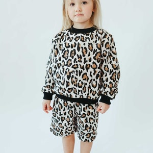 May include: A child's two-piece set featuring a leopard print design. The set includes a long-sleeved sweatshirt and matching shorts. The sweatshirt has black trim at the neckline and cuffs. The shorts have a similar black trim.