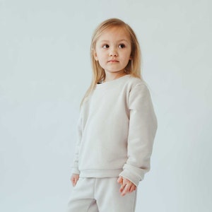 Pode incluir: Uma criança pequena vestindo um moletom bege claro e shorts combinando. A criança está de pé com os pés descalços e olhando para o lado.