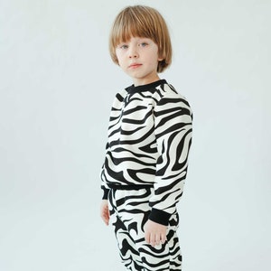 May include: A young person wearing a black and white zebra print sweatshirt and matching pants. The sweatshirt has a round neckline and long sleeves. The pants have a drawstring waistband and a relaxed fit.