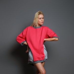 May include: A woman wearing a pink and gray oversized sweatshirt with a gray trim. She is wearing white sneakers and standing on a gray background.