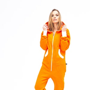 Może przedstawiać: Pomarańczowy kombinezon z kapturem i białymi lamówkami wokół kaptura, mankietów i kieszeni. Kombinezon zapinany na zamek błyskawiczny, o luźnym kroju.