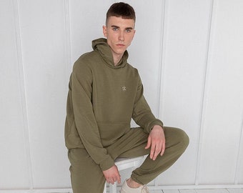 Könnte beinhalten: Ein grüner Kapuzenpullover und eine passende Jogginghose mit einem kleinen gestickten Logo auf der Brust. Das Model trägt das Outfit und sitzt auf einem weißen Hocker.