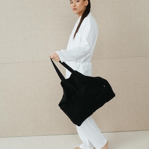 May include: A person wearing a white jumpsuit holds a black tote bag. The bag has a wide, flat base and two long straps.