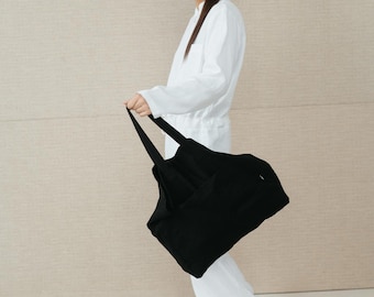 May include: A person wearing a white jumpsuit holds a black tote bag. The bag has a wide, flat base and two long straps.