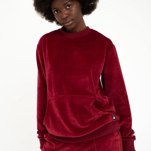 K&ouml;nnte beinhalten: Ein weinroter Samt-Sweatshirt mit einer K&auml;ngurutasche und einer passenden Hose. Der Pullover hat ein kleines schwarzes Logo unten rechts.