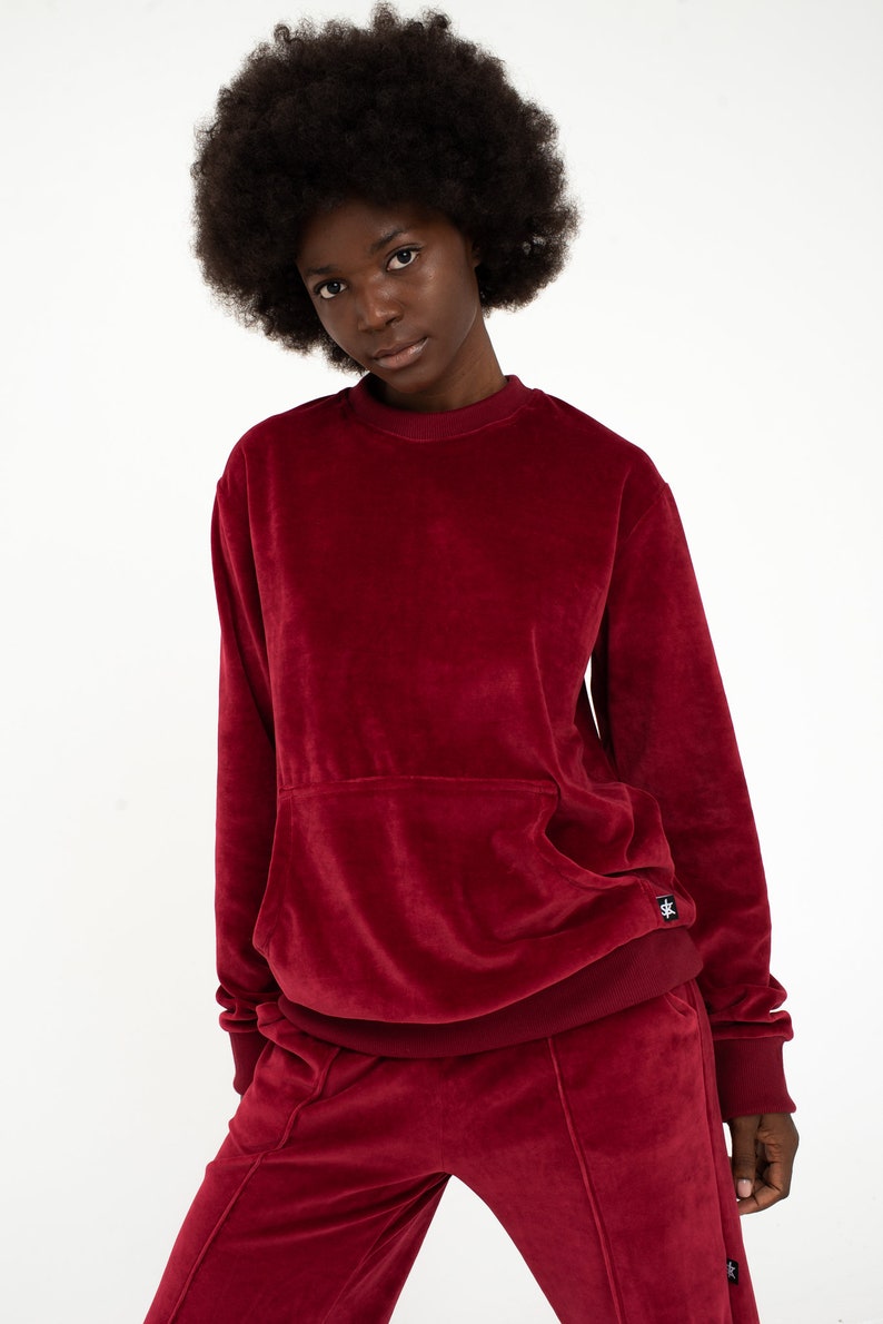 K&ouml;nnte beinhalten: Ein weinroter Samt-Sweatshirt mit einer K&auml;ngurutasche und einer passenden Hose. Der Pullover hat ein kleines schwarzes Logo unten rechts.