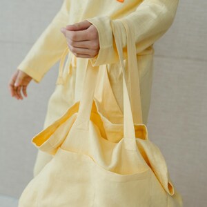 May include: A pale yellow linen tote bag with a large pocket on the front. The bag has two long straps and a small black tag with a logo on the bottom right corner.