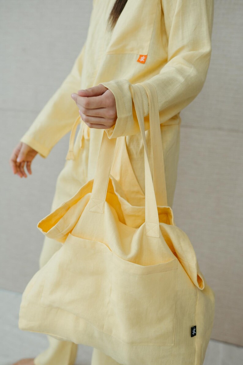 May include: A pale yellow linen tote bag with a large pocket on the front. The bag has two long straps and a small black tag with a logo on the bottom right corner.