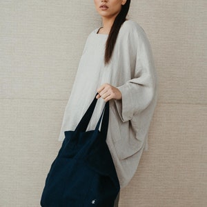 May include: A woman wearing a light gray linen top and gray pants is holding a dark blue tote bag with a small white logo.
