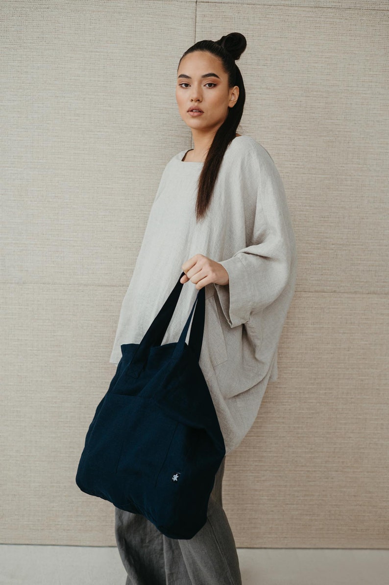 May include: A woman wearing a light gray linen top and gray pants is holding a dark blue tote bag with a small white logo.